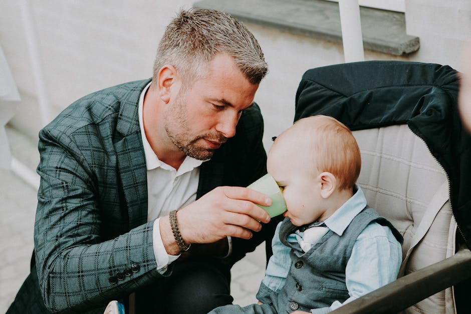Concentrated adult father in white shirt and jacket giving toddler in elegant clothing water in cup on street in daylight
